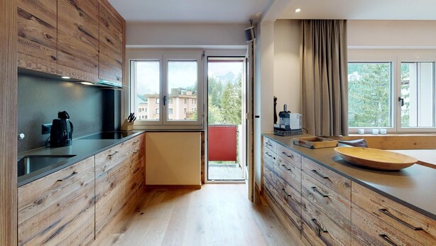 A modern kitchen with wooden cabinetry, a built-in oven, and a large window offering a view of buildings and trees. - Apartmenthouse house next to the lake in St. (St. Moritz)