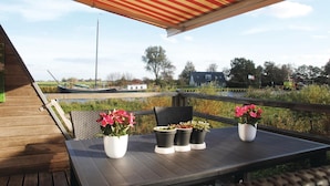 A wooden deck with a table and chairs, potted plants, and a view of boats and houses across a water body.