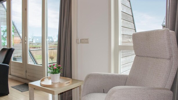 A room with a large window, a grey armchair, a small table with a white vase and pink flowers, and a black chair.