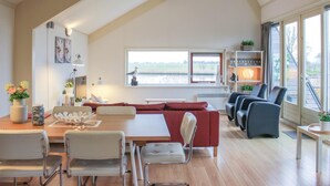 A spacious living area with a dining table, chairs, a red sofa, and a large window overlooking a body of water.