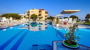 A swimming pool with lounge chairs and umbrellas, surrounded by a building with multiple floors and balconies. - Matzi Apartments (Platanias)