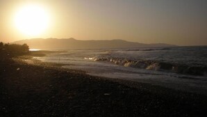 A beach scene with the sun setting over the ocean, waves gently crashing onto the shore, and distant mountains in the background. - Matzi Apartments (Platanias)