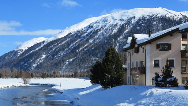 A snow-covered landscape with a building featuring balconies and a river. - The traditional Chesa Marmotta is situated next to the famous River "Inn" in a v (Celerina/Schlarigna)
