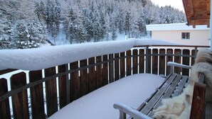 Un balcón de madera con barandilla de metal, una banca y un perro sentado en ella. Nieve cubre el suelo y los árboles al fondo.