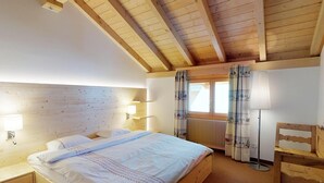 Un dormitorio con techo de madera, una cama con ropa de cama blanca, una mesita de noche, una lámpara de pie y una ventana con cortinas.