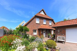 A two-story brick house with a red-tiled roof, a front porch, and a garden with various plants and flowers. - Apartment 1 - House Strandlooper (Neuharlingersiel)