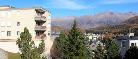 Un edificio de varios pisos con balcones, rodeado de árboles de hoja perenne y montañas al fondo.