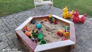 Ein Sandkasten mit Spielzeug, eine Bank und ein Garten im Hintergrund.