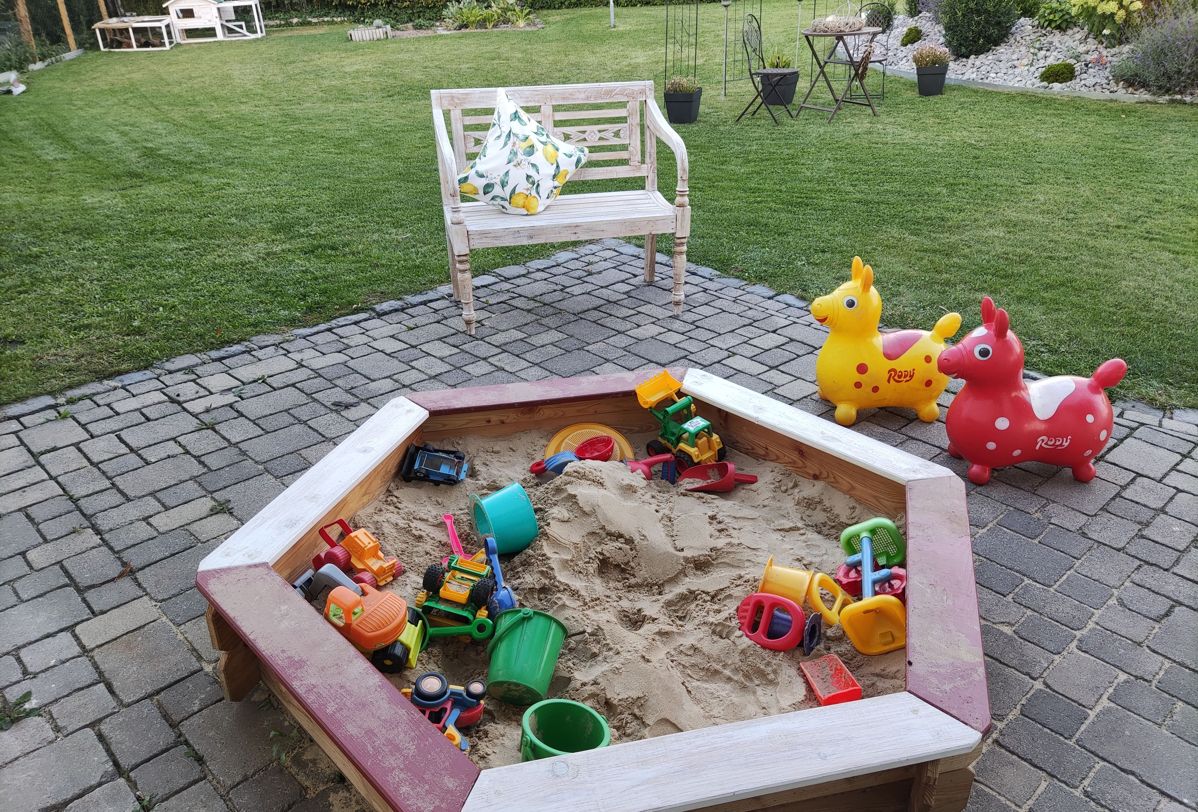 Ein Sandkasten mit Spielzeug, eine Bank und ein Garten im Hintergrund.