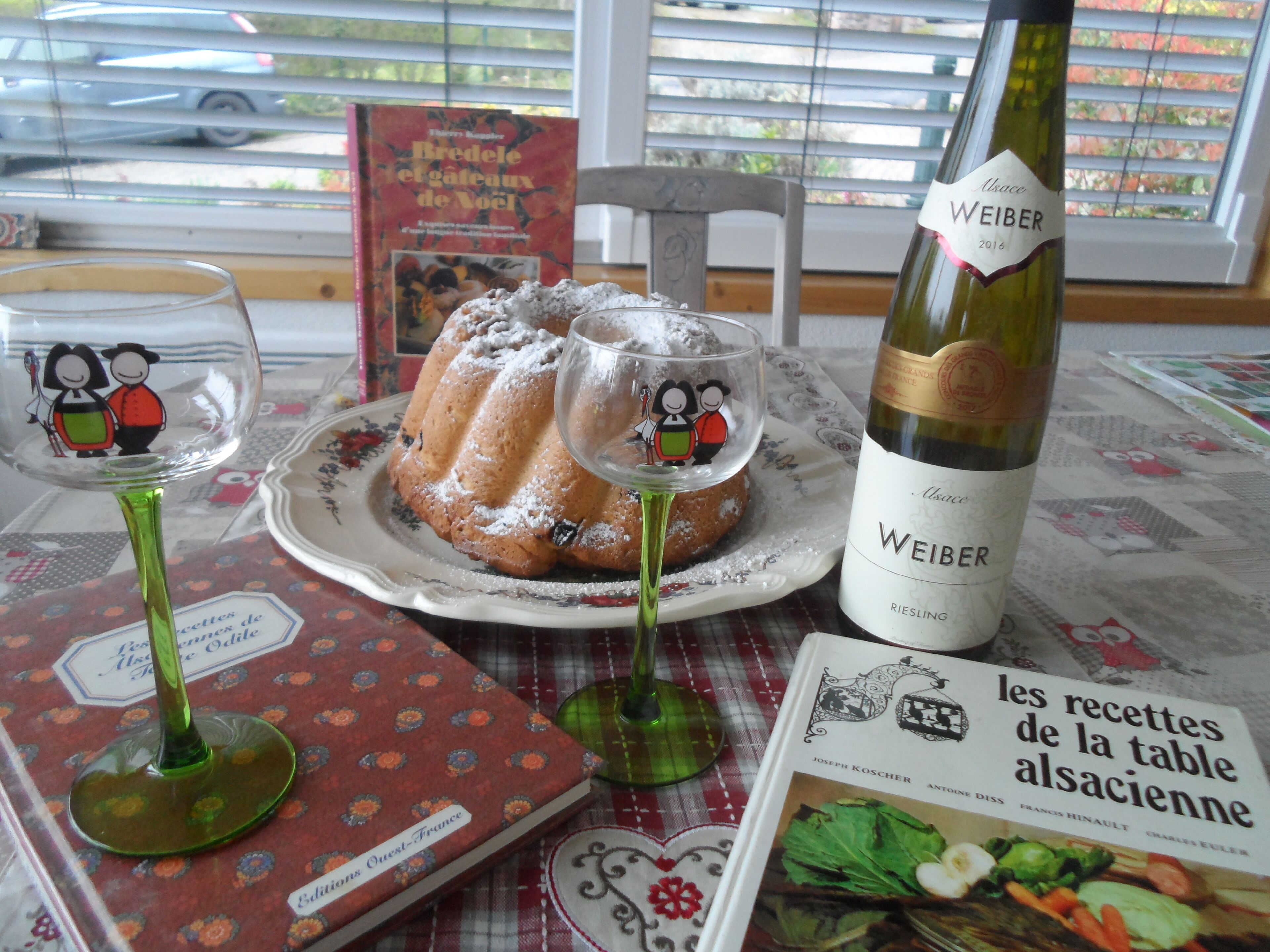 A table with a bundt cake, a bottle of Riesling wine, and a cookbook titled 'les recettes de la table alsacienne'.