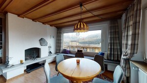 Ein gemütliches Wohnzimmer mit Kamin, einem runden Holztisch und Blick auf Berge durch große Fenster.