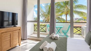 Une chambre d’hôtel avec un lit, un téléviseur posé sur une commode en bois, et un balcon offrant une vue sur les palmiers et la mer.
