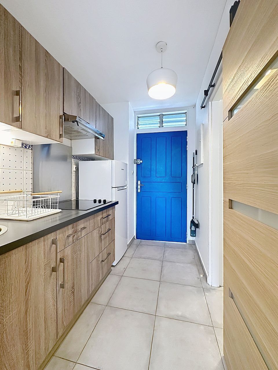 Une cuisine avec des meubles de rangement en bois, un plan de travail blanc, une porte bleue et un luminaire au plafond.
