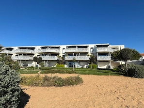 A modern building with multiple balconies, a sandy foreground, and greenery.