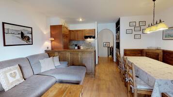 Ein gemütliches Wohnzimmer mit grauem Sofa, Holztisch und einem Essbereich mit Tisch und Stühlen. Im Hintergrund ist eine Küche mit Holzausstattung und gefliestem Rücksprühefeld zu sehen.
