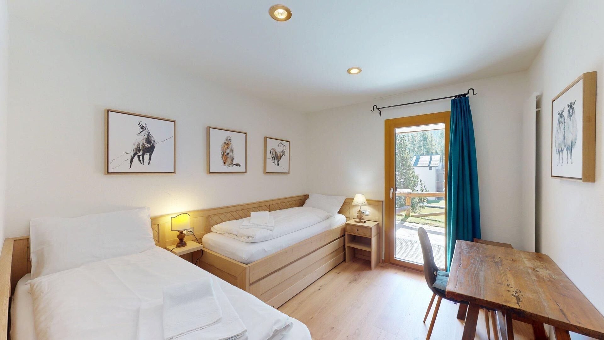 Ein Zimmer mit zwei Betten, einem Holztisch und einem Stuhl. Drei gerahmte Bilder hängen an der Wand.