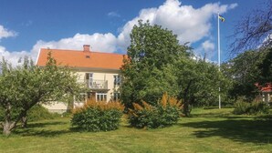 Een huis met twee verdiepingen, met een dak van rode tegels, omgeven door weelderige begroeiing en een helderblauwe lucht.