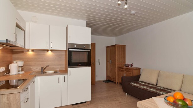 A compact kitchen with white cabinets, a built-in oven, and a sink. There's a wooden dining table with a bowl of fruit and a coffee maker. - 02 Apartment Sonnleiten 35 sqm for max. 3 p., kitchen, 1 living/bedroom, 1 bedroom, balcony (Bergen)