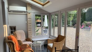Ein sonnendurchfluteter Raum mit Korbmöbeln, einem Glastisch und Blick auf einen Garten durch große Fenster.