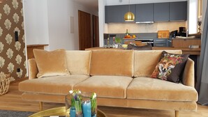 Ein modernes Wohnzimmer mit einem beigefarbenen Sofa, einem Glastisch mit Blumen und einer Küche im Hintergrund.