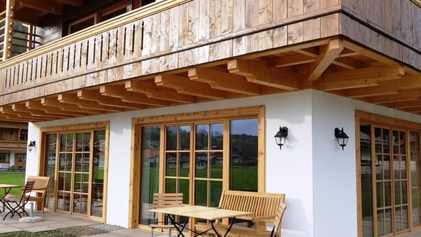 Ein zweistöckiges Holzhaus mit Balkon, großen Fenstern und Außenbereich mit Sitzgelegenheiten.