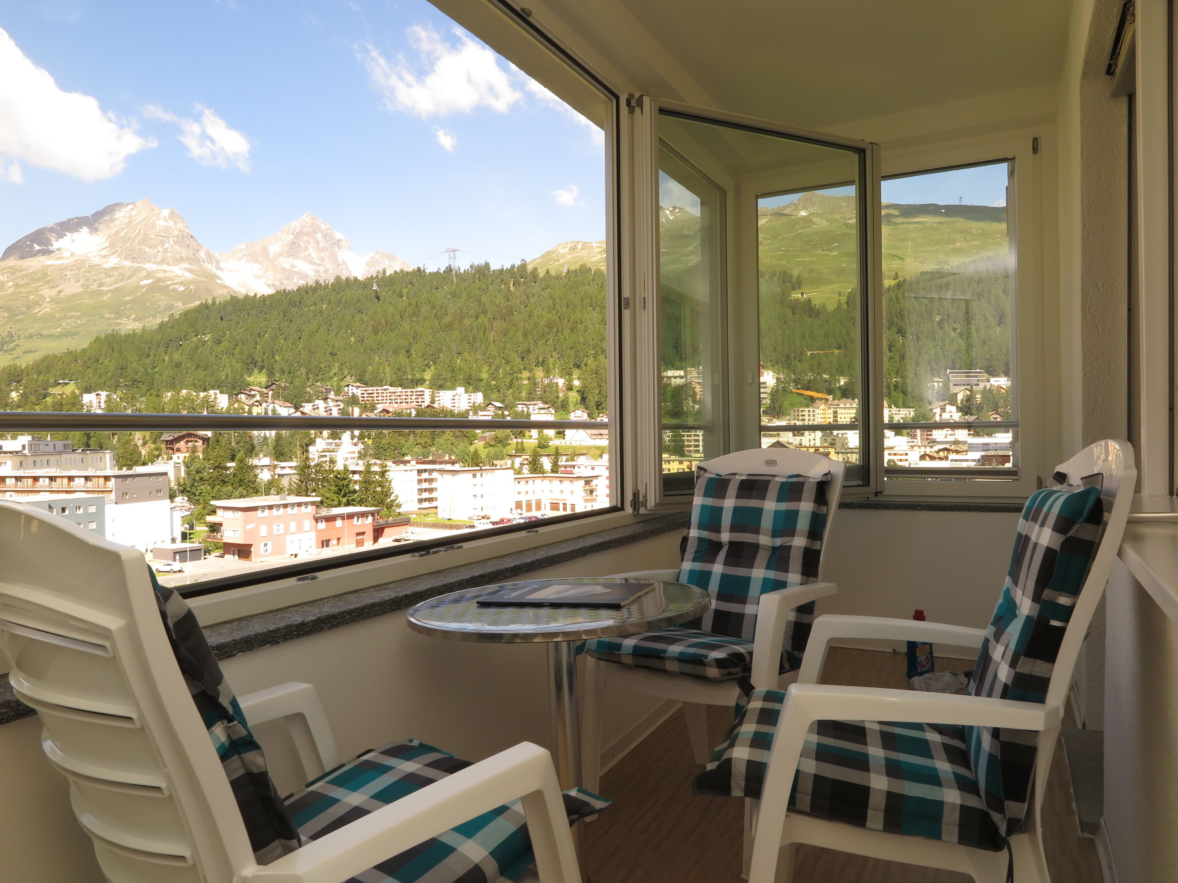 Un balcon avec deux chaises et une petite table, d’où l’on peut admirer un paysage montagneux et une ville.