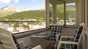 A balcony with two chairs and a small table, overlooking a mountainous landscape and a town. - Apartment building with excellent infrastructure. (St. Moritz)