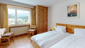 A bedroom with a wooden bed, a chair, a desk, a window with curtains, and a painting on the wall. - Apartment building with excellent infrastructure. (St. Moritz)
