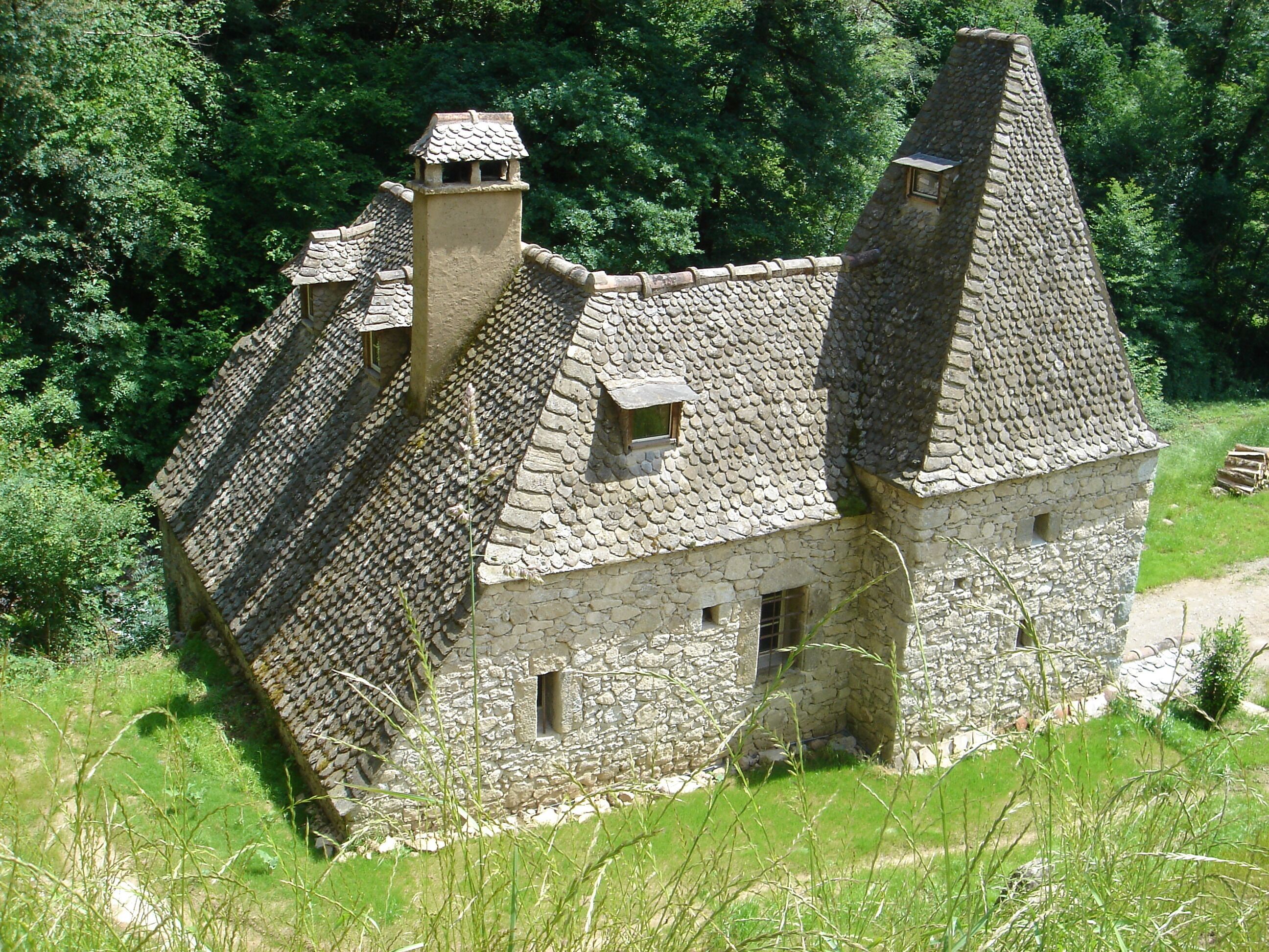 Un bâtiment en pierre, doté de toits à forte pente et de nombreuses cheminées, entouré d’une végétation luxuriante.