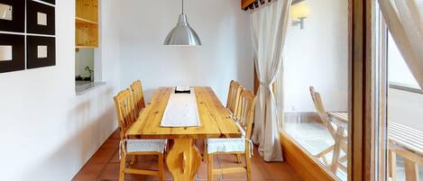 A dining area with a wooden table and chairs, a pendant light, and a view through a window with curtains.