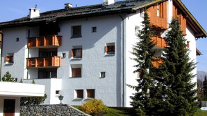 Un edificio de varios pisos con balcones, un muro de piedra y un gran árbol de hoja perenne.