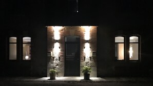 Un bâtiment la nuit, avec des fenêtres illuminées et une porte, ainsi que des plantes en pot devant l’entrée.