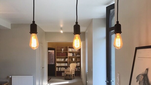 Un couloir avec trois luminaires suspendus, un radiateur, une bibliothèque et un tableau encadré accroché au mur.