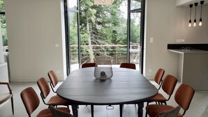 Une salle à manger moderne avec une table ronde entourée de chaises, de grandes fenêtres donnant sur la verdure et un coin cuisine agrémenté de luminaires suspendus.