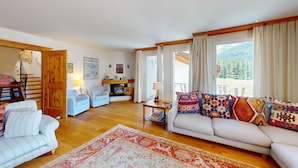 Un salon chaleureux avec un canapé, deux fauteuils, une table basse en bois et un tapis à motifs. Un escalier mène à un niveau supérieur, et une grande fenêtre offre une vue sur une zone boisée.