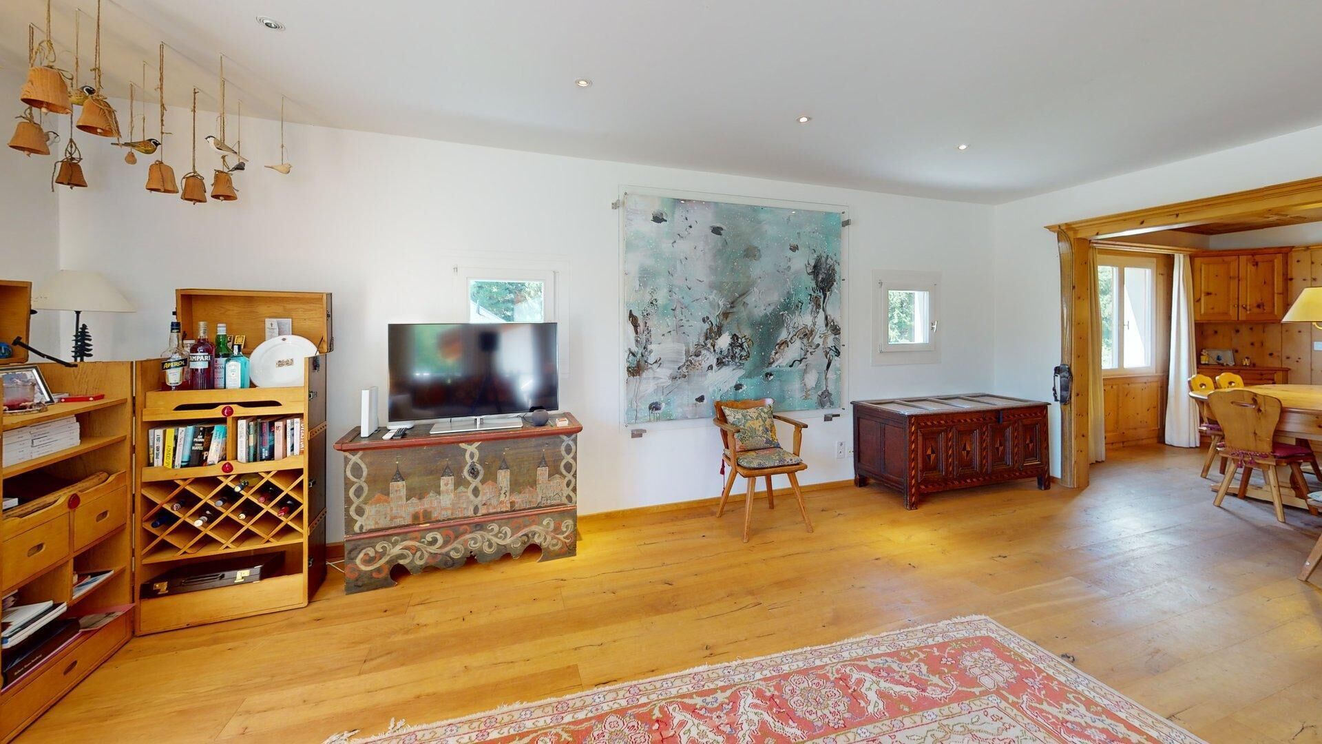 A spacious living room with a wooden floor, a large painting on the wall, a television, and a dining area with wooden furniture.