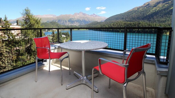 A balcony with two red chairs and a round table, overlooking a turquoise lake and mountains. - Terraced apartmentbuilding between St. (St. Moritz)