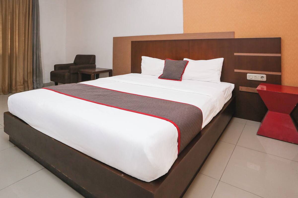 A hotel room with a large bed, a wooden headboard, a red side table, and a chair.