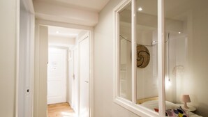 Un couloir étroit, doté d’un plancher en bois, mène à une pièce avec une grande fenêtre, un lit et une table de chevet avec une lampe.
