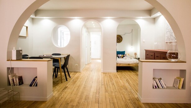 Une pièce compacte et moderne, dotée d’un plancher en bois, d’un coin repas avec une table et des chaises, d’un lit avec une tête de lit et d’une petite armoire contenant des livres.
