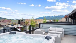 Una terraza en la azotea con jacuzzi, muebles de mimbre y vistas a un pueblo con montañas al fondo.