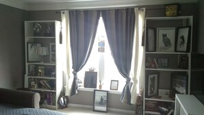 A room with a large window, curtains, a bookshelf, and a bed. - CrownCity Home Away From Home (Charlotte)
