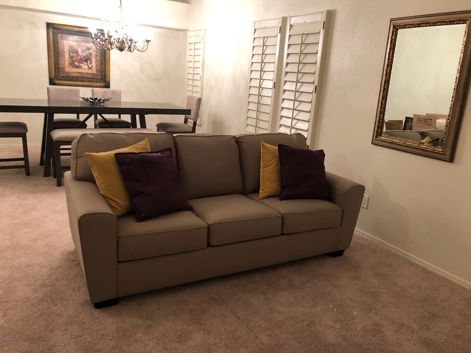 A living room with a beige sofa, a dining table with chairs, a chandelier, and a mirror on the wall.