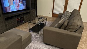 A living room with a television on a wooden stand, a glass coffee table, and a brown sofa with patterned cushions. - Very nice 4 bdrm 2 full bath ranch home w/ great room in quiet neighborhood. (Phoenix)