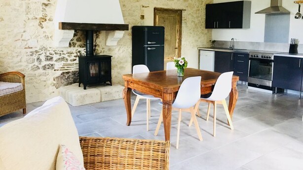 A rustic dining area with a wooden table and chairs, a stone wall, a wood-burning stove, and a modern kitchen in the background. - Beautifully restored 2 bedroom house with private pool and barbecue. (Thénac)