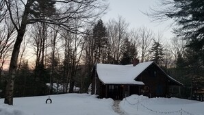 A rustic cabin with a snow-covered roof, surrounded by trees and a swing set in the foreground. - Private Brook Side Log Cabin  on Tenney Mountain (Plymouth)