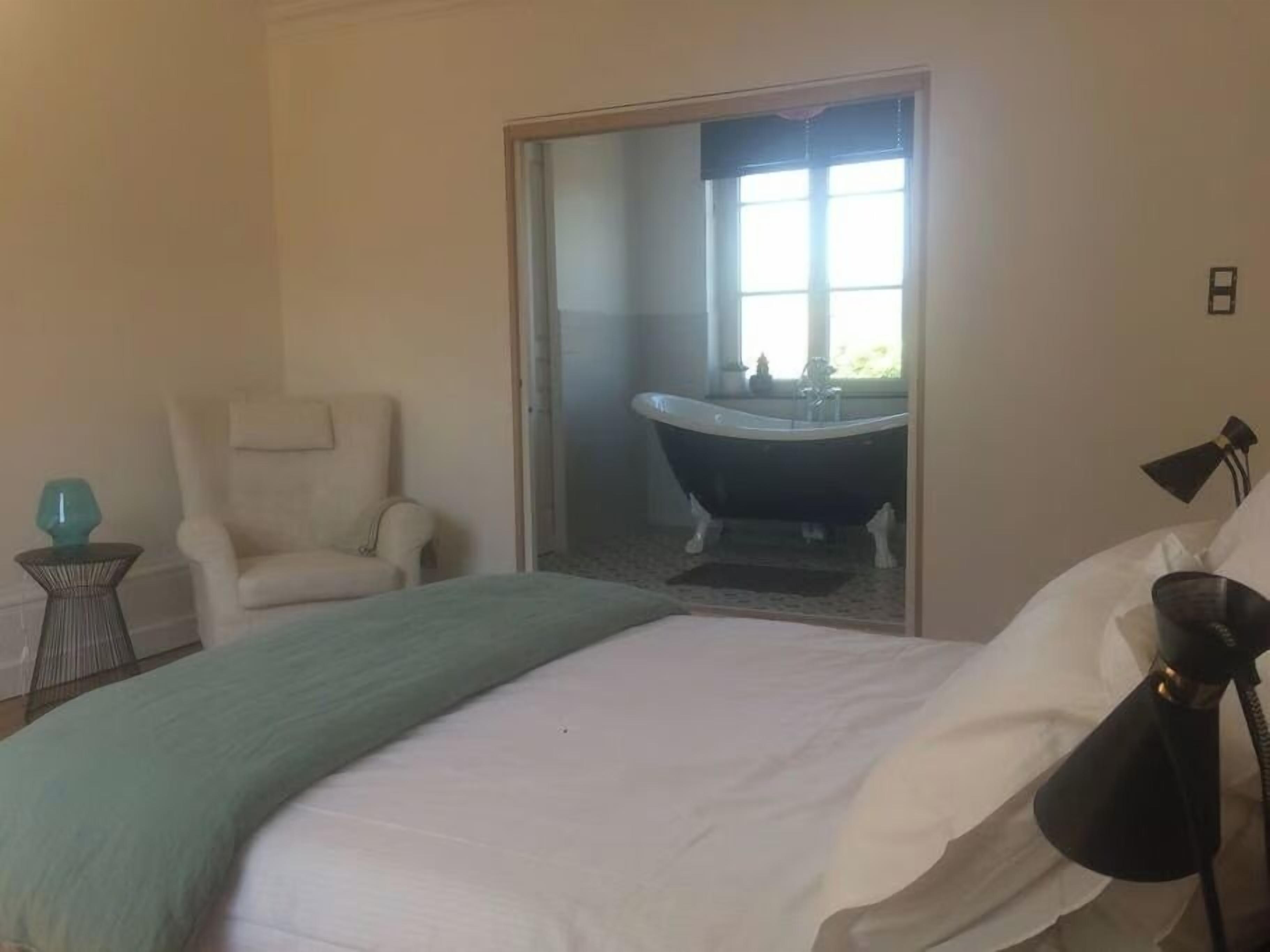A bedroom with a bed, a chair, a bedside table, and a view of a bathroom with a bathtub.
