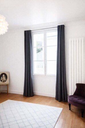 Ein Zimmer mit großem Fenster, dunklen Vorhängen, einem violetten Sessel und einem gerahmten Bild an der Wand.