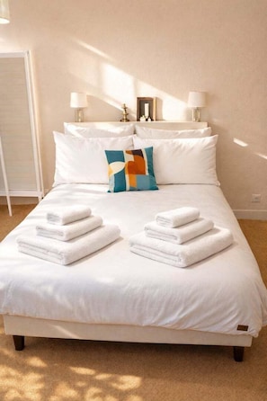 Een net opgemaakt bed met witte lakens en opgevouwen handdoeken, een kleurrijk kussen en twee bedlampen op een witte nachtkastje.