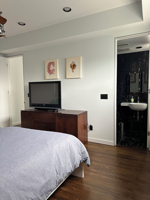 A bedroom with a bed, a TV on a wooden dresser, and a bathroom with a sink and mirror.
